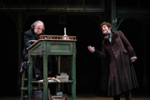 Scrooge sits at a desk while a young man speaks with him in A Christmas Carol