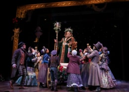 The Ghost of Christmas Present stands on a sleigh above a group of people in A Christmas Carol