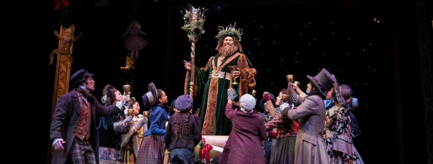 The Ghost of Christmas Present stands on a sleigh above a group of people in A Christmas Carol