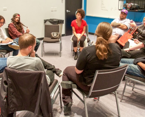 A group of actors sit in a circle in a studio, some holding notepads and paper