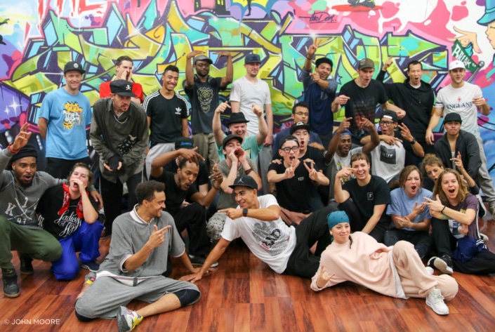 A large troupe of dancers poses for a group shot in front of a wall of graffiti art