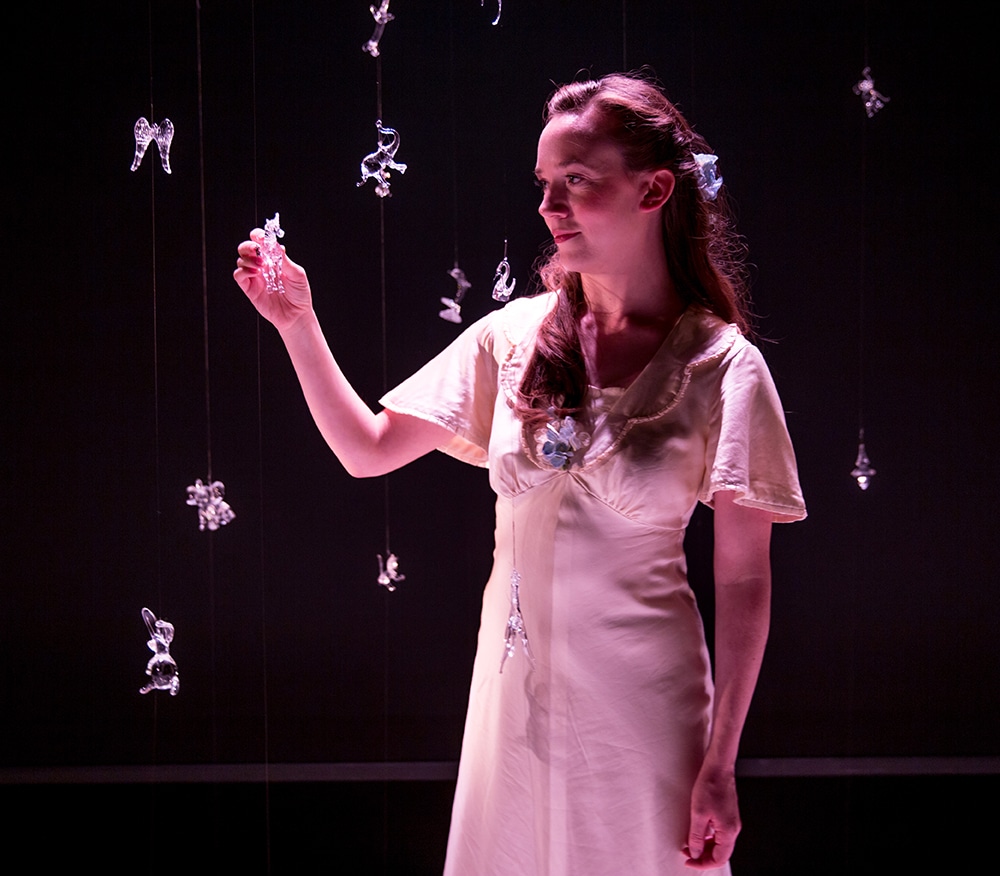 Woman in a 40s style white gown stares at the small floating glass trinkets around her in a production of The Glass Menagerie