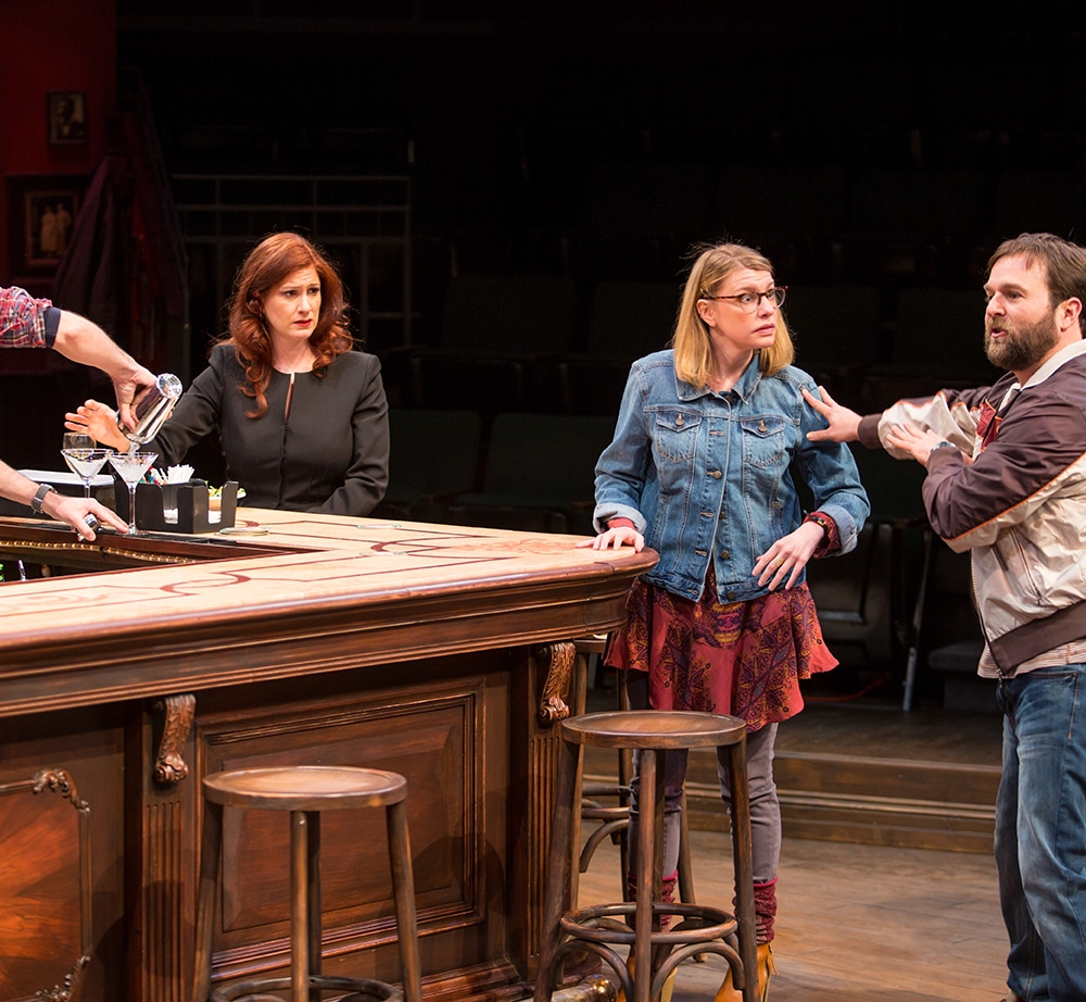 Two women in a pub at the bar stare in shock as the man on the right shares some news in a production of "The Nest"