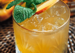 a rocks glass filled with a yellow beverage and topped with a peach slice and mint leaves is sitting in a brown wicker surface with whole peaches in the background