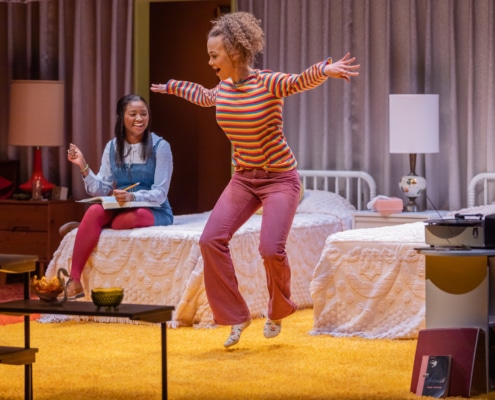 An actor sits on a bed watching as another actor dances in In the Upper Room.