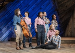 Seven female cast members, including Pekarek, pose and sing on a stage made of distressed wood in front of a blue backlight