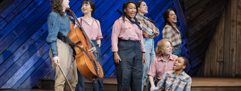 Seven female cast members, including Pekarek, pose and sing on a stage made of distressed wood in front of a blue backlight