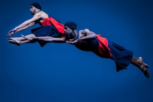 CPRD Ensemble dancers leap in front of a blue background wearing black and red costumes