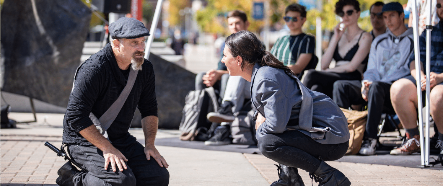 Actor in silver sash and newsboy cap on his knees conversing with actress in a silver blazer and sash crouching to his level