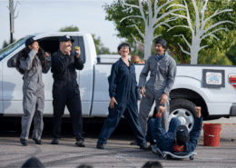 In front of a white pickup truck, ensemble of actors in carpenter suits cheer on actress in a newsboy cap