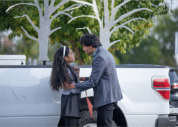 Actor in a silvery blue blazer holds Actress in a white sweater and headband as they exchange deep and heart felt looks