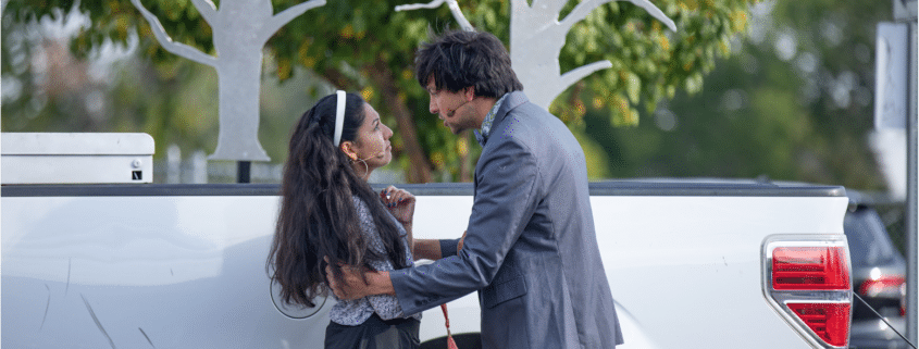 Actor in a silvery blue blazer holds Actress in a white sweater and headband as they exchange deep and heart felt looks