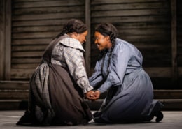 Two women kneel, facing one another with their hands clasped. They are both smiling brightly.