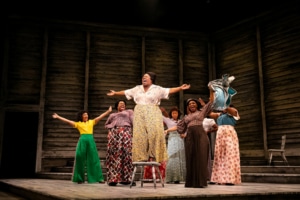 Several women sing onstage, clustered together with their arms outstretched. The woman in front is standing on a chair as she belts out a note.