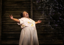 A woman stands with her arms outstretched, belting a song onstage.