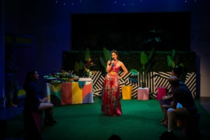 Maggie Bofill holds one hand on her hip and a microphone in the other while standing in the middle of a colorful courtyard. There is an elaborate spread of food on a table behind her, and Stephanie Machado, Danielle Alonzo, and Luis Vega sit watching her.