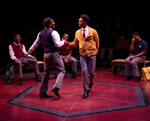 Two males are dancing mid stage in front of three other school mates sitting on the benches behind