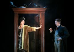 Henry Walter Greenberg dressed as Mrs. Jordan in a party dress stands in the doorway holding a handout to stop Richard Hannay