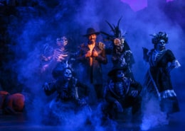 A man in a black coat is surrounded by Calacas and blue smoke