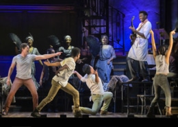 A man standing on a table plays guitar while other cast members react in Hadestown