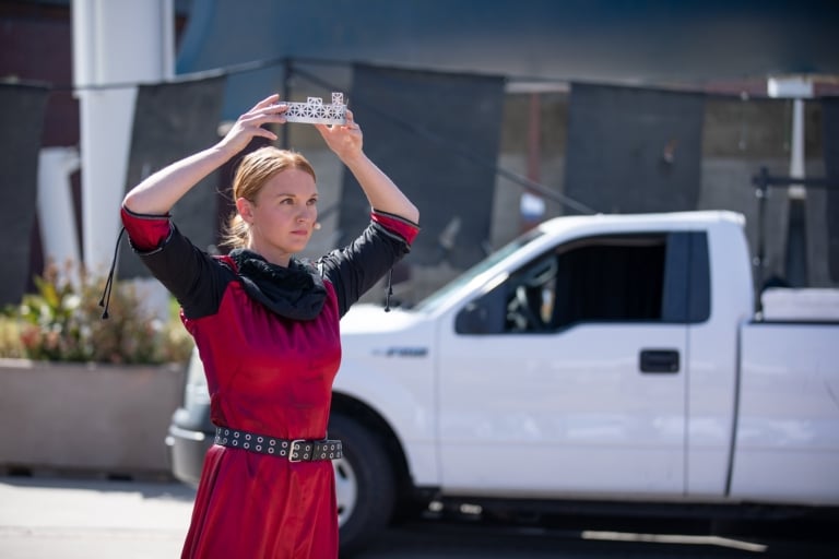 Female actor with blond hair in red costume dress holding a metal crown above her head with a serious expression on her face.