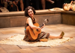 A woman sits onstage with a guitar in Much Ado About Nothing