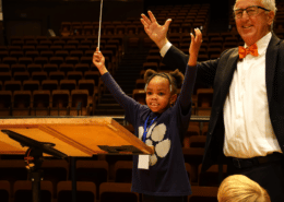 A young girl stands next to a conductor, both with the arms in the air