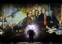 A woman stands onstage in front of Revolutionary War artwork in 1776