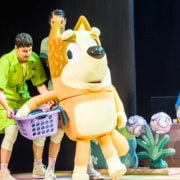 Mum is carrying a laundry basket, supported by actors in green jumpsuits.