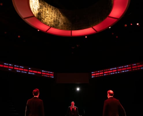 Three men in black suits and coats face each other. They are washed in red light from the chandelier above them. There is ticker tape similar to what is on the New York Stock Exchange on the wall behind them.
