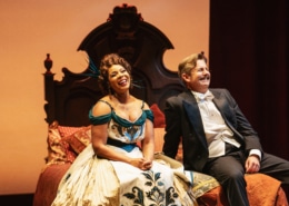 Soara-Joy Ross and Edward Staudenmayer laugh on an intricate bed in elegant costumes.