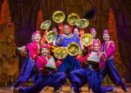 Genie points to the audience surrounded by a group of colorfully dressed dancers. The dancers show off desserts from beneath golden cloches