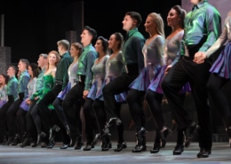 A row of smiling dancers wearing green and black costumes performing in Riverdance