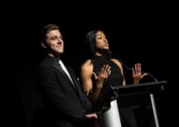 Thomas Beeker and Madison Manning, wearing black tie, present at a podium during the 2023 Bobby G Awards