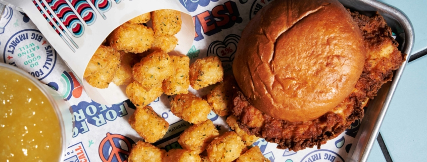 A fried chicken sandwich with a pouch of tater tots spilling on a Birdcall tray