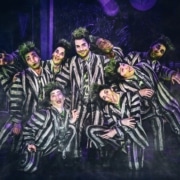 Beetlejuice, wearing his signature black and white striped suit, is surrounded by matching dancers imitating his insane expression