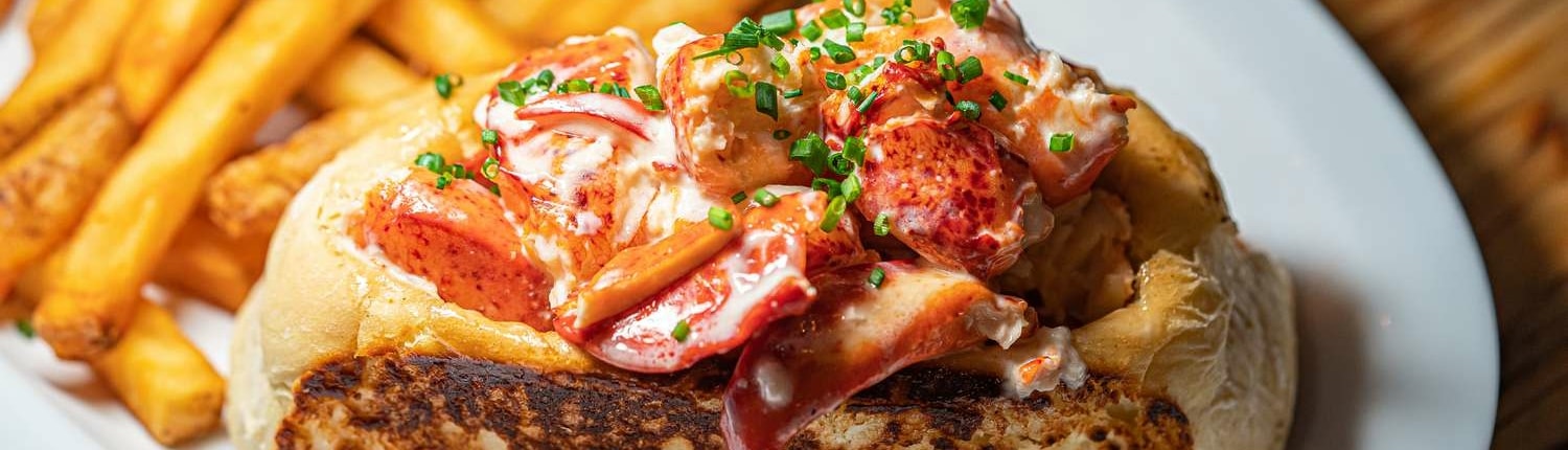 A fresh lobster roll topped with chopped herbs. Served on a plate with fries.