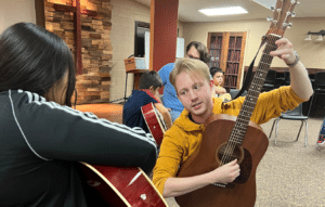 Two teens sitting across from each other observe the other's guitar skills