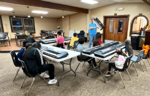 Kids sit at long folding tables, watching a teacher holding a small keyboard