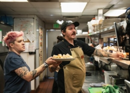 A man hands plates filled with food over to a woman. They are in the kitchen of Steuben's