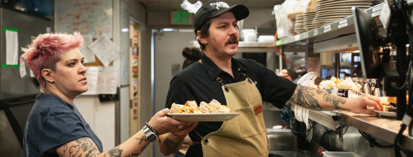 A man hands plates filled with food over to a woman. They are in the kitchen of Steuben's
