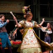 A man claps while a woman dances, her skirts flying. A man with a violin plays in the background.