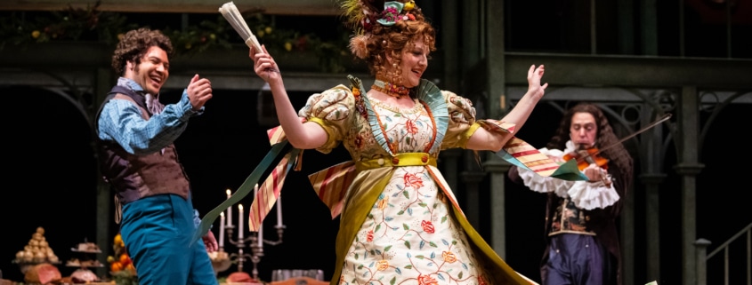 A man claps while a woman dances, her skirts flying. A man with a violin plays in the background.