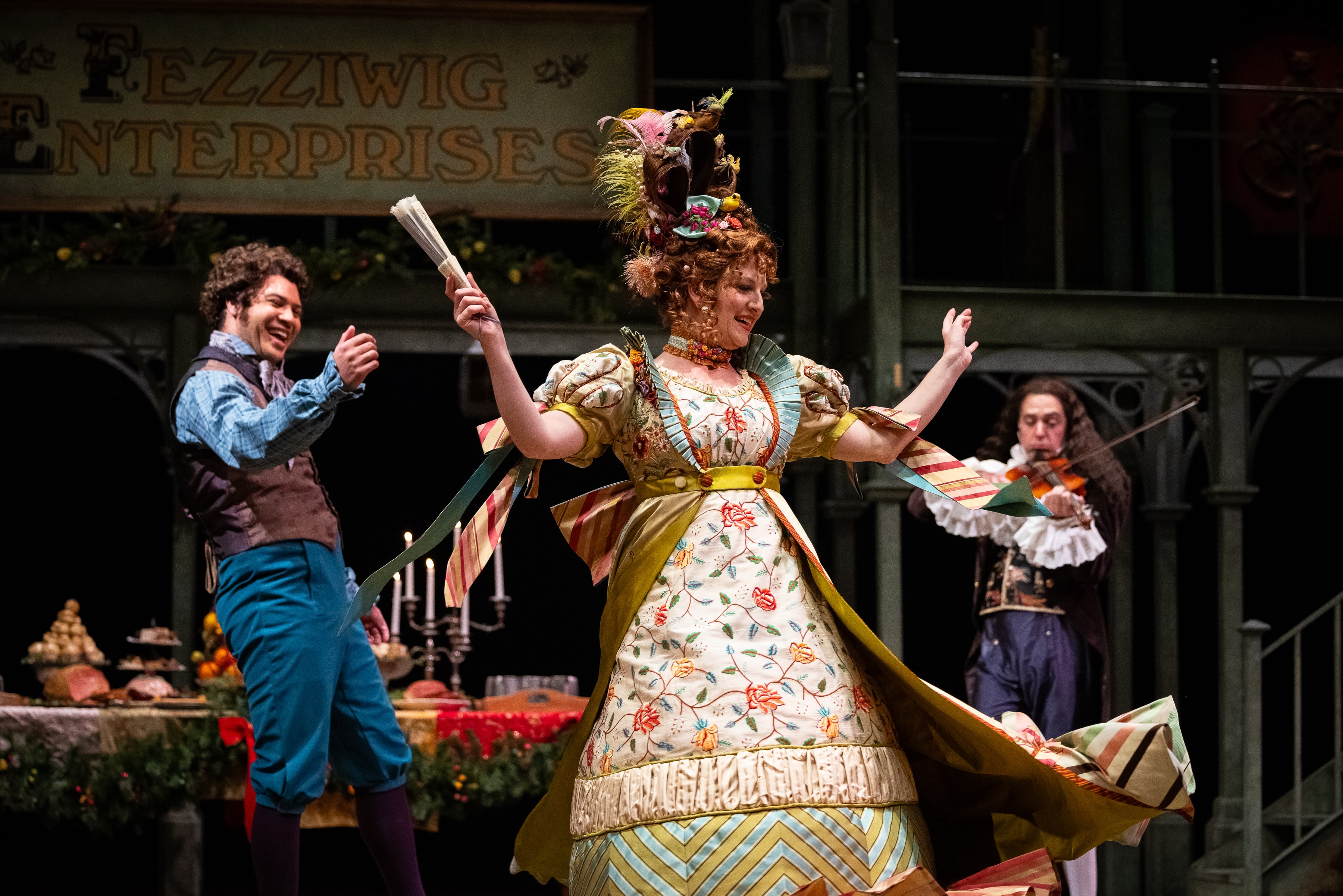 A man claps while a woman dances, her skirts flying. A man with a violin plays in the background.