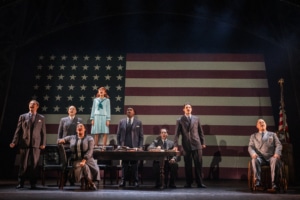 A young girl stands on a table to sing, surrounded by older men in suits. They are all posed in front of a large American flag.