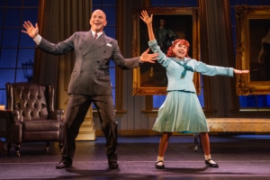 A man and young girl dance in a formal living room. She is wearing a sailor style dress, he is in a suit and tie.