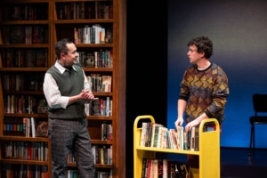 A man in a green sweater vest clasps his hand, while talking to a young man in a colorful sweater. They are in a bookstore the young man is shelving books.