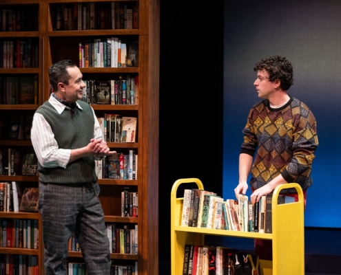 A man in a green sweater vest clasps his hand, while talking to a young man in a colorful sweater. They are in a bookstore the young man is shelving books.