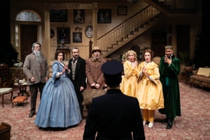 A group of people in 19th century clothes gasp dramatically as they hear news from the constable who is facing away from the camera.