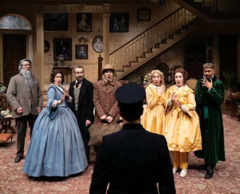 A group of people in 19th century clothes gasp dramatically as they hear news from the constable who is facing away from the camera.